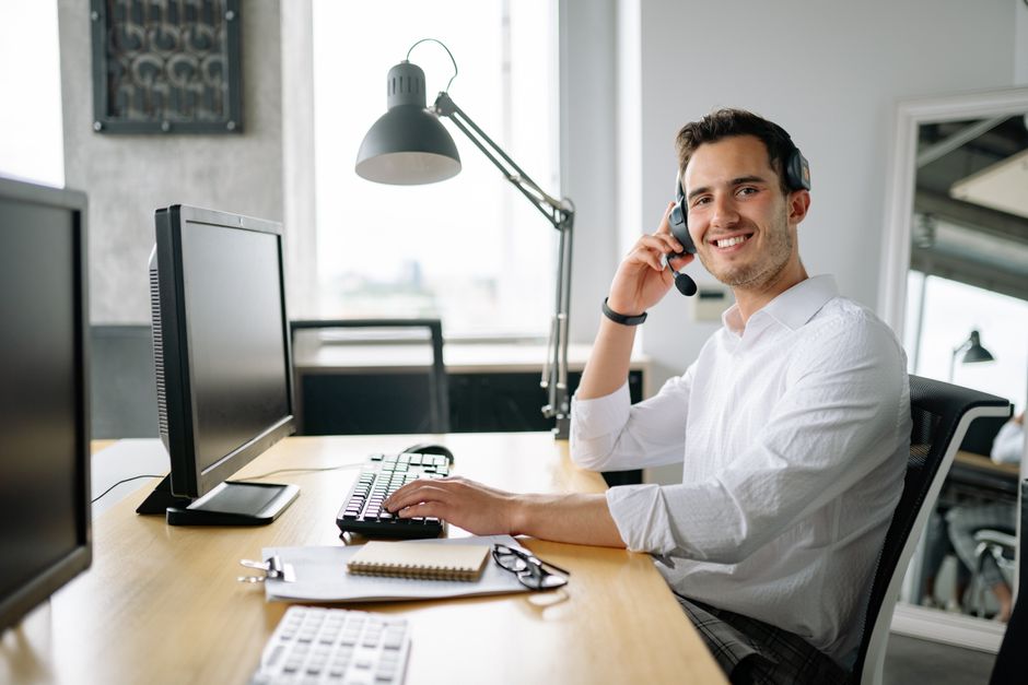 Agente de call center sorridente trabalhando em uma mesa com um headset e computador em um escritório moderno.