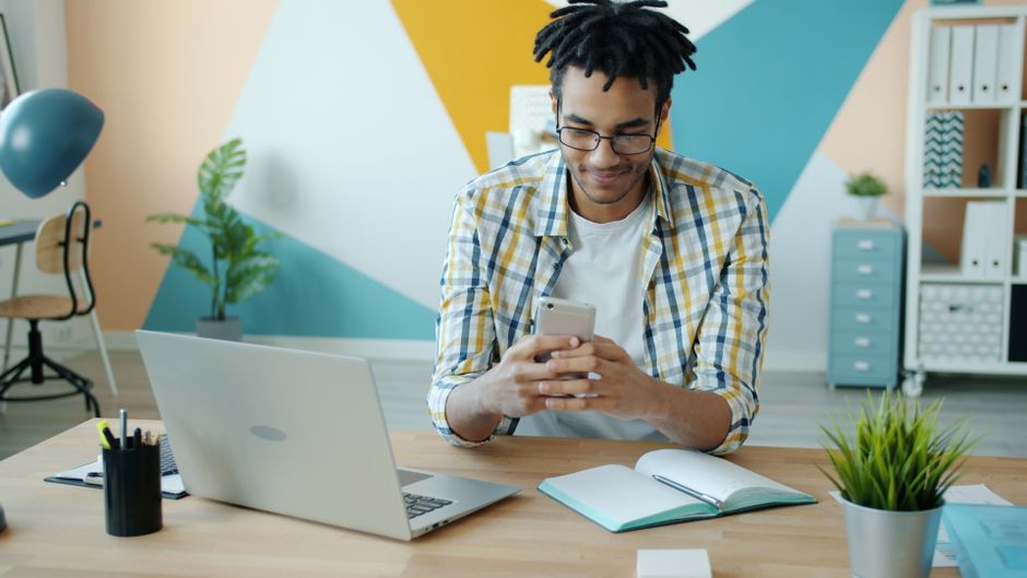 Jovem adulto usando um smartphone em uma mesa com um laptop em um escritório moderno.