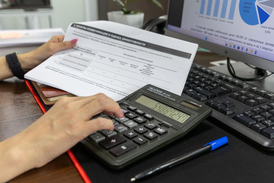 Uma pessoa calcula dados financeiros usando uma calculadora e um documento, trabalhando em uma mesa de escritório.