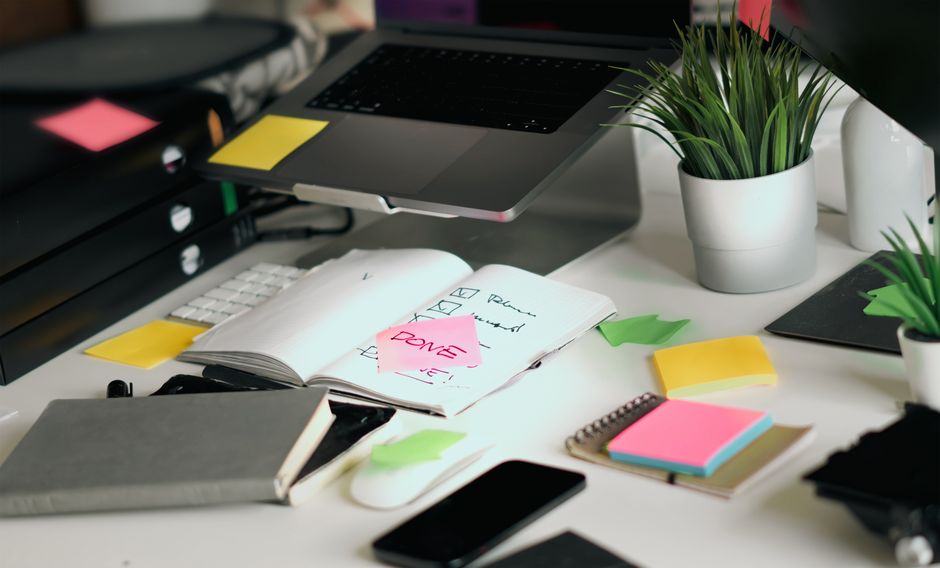 Uma mesa de escritório desorganizada com um laptop, cadernos e listas de verificação em meio a post-its coloridos.